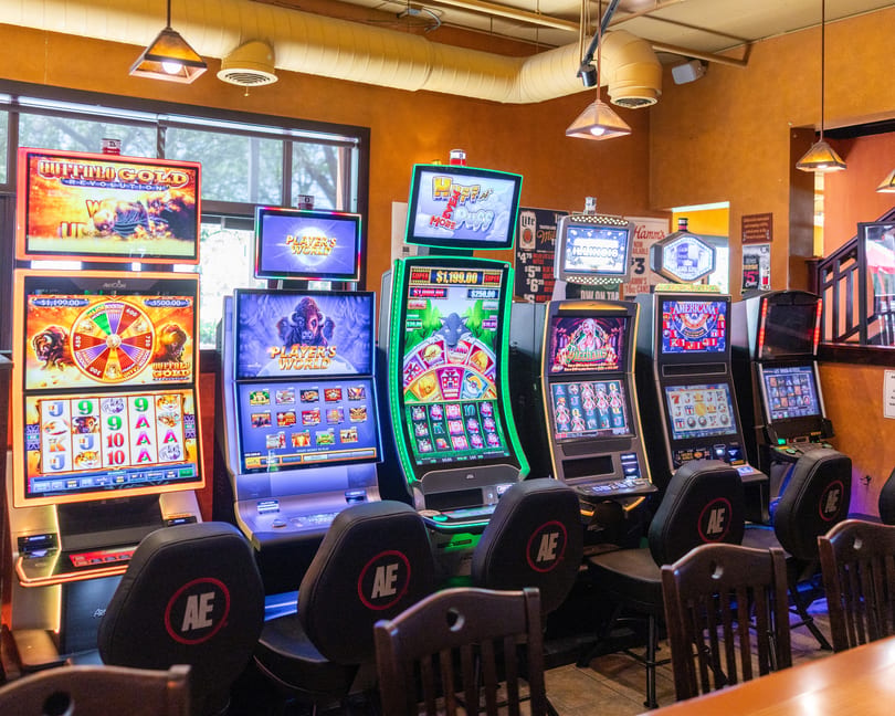 A row of brightly lit slot machines with various games and chairs in a casino or bar.