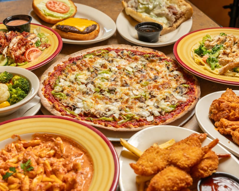 A table laden with various dishes including pizza, pasta, burgers, salads, and fried shrimp.