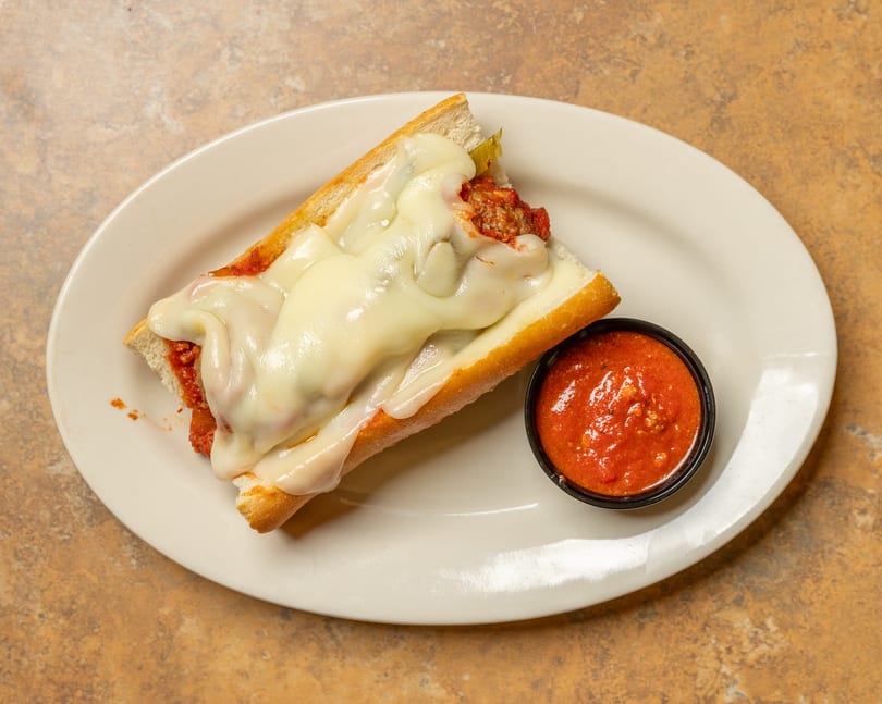 Meatball sub sandwich with melted mozzarella cheese and a side of marinara sauce on a white plate.